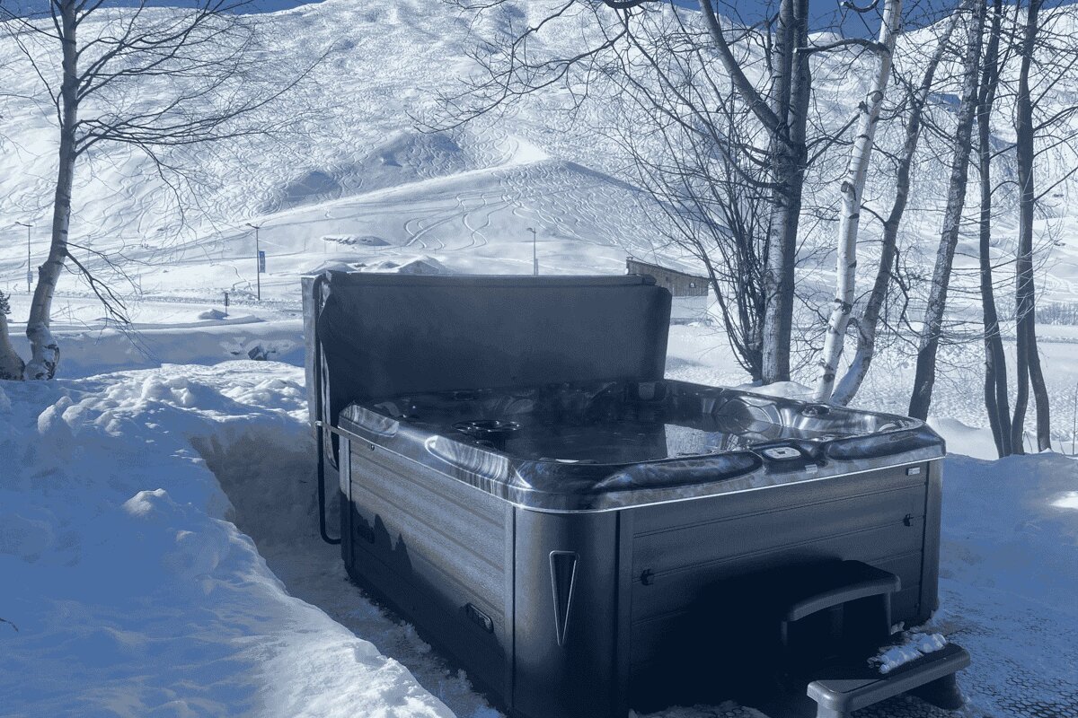 An outdoor hot tub sits open in a snowy landscape, surrounded by bare trees with snow-covered mountains in the background.