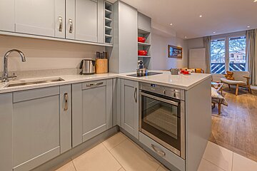 A modern light grey kitchen with an integrated oven and sink, leading to an open-plan living area with a large window overlooking a street.