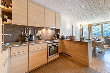 A modern kitchen with light wood cabinets, stainless steel appliances, and a dining area overlooking a scenic forest view.