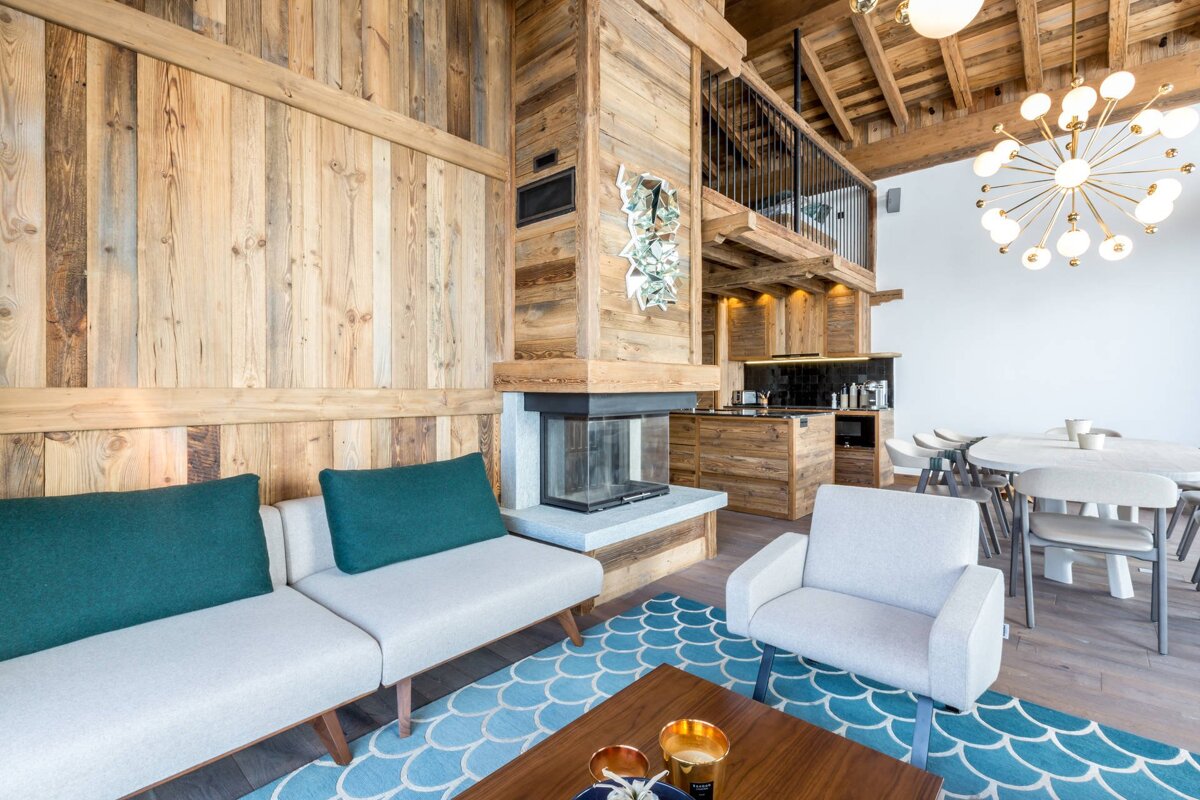 Modern chalet interior featuring wood-paneled walls, a fireplace, contemporary furniture, a blue patterned rug, and a dining space.