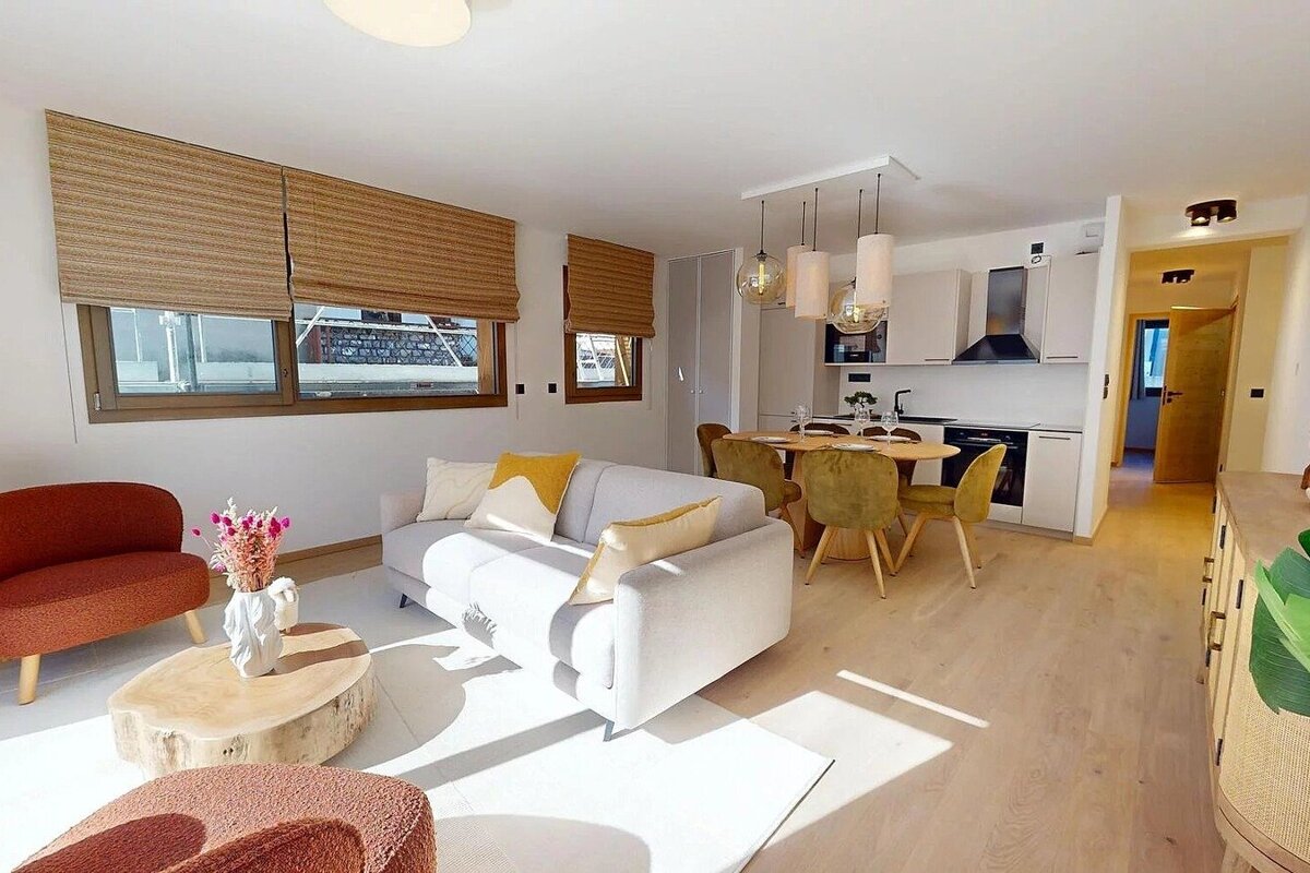 A modern, sunlit open-plan living room and kitchen featuring light wood floors, a gray sofa, coral accents, and a dining area.