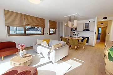 A modern, sunlit open-plan living room and kitchen featuring light wood floors, a gray sofa, coral accents, and a dining area.