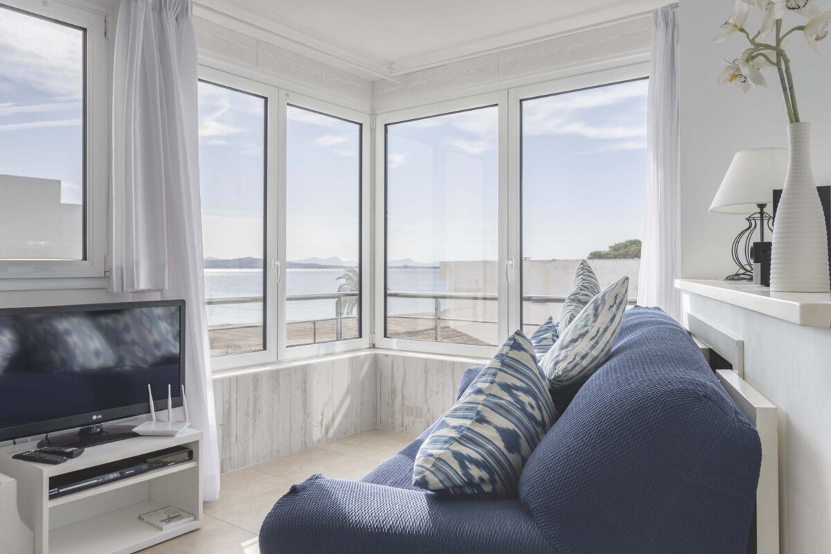 A cozy living room with a blue sofa, large windows showcasing a beautiful ocean view, a TV, and white decor.