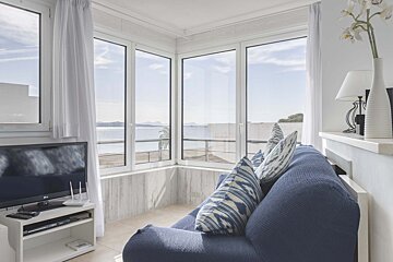 A cozy living room with a blue sofa, large windows showcasing a beautiful ocean view, a TV, and white decor.
