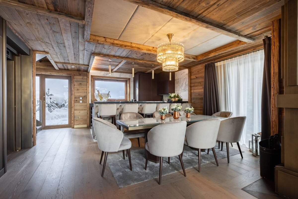 A luxurious ski chalet dining area with rustic wood, elegant velvet chairs, a modern chandelier, and a snowy outdoor view through the window.