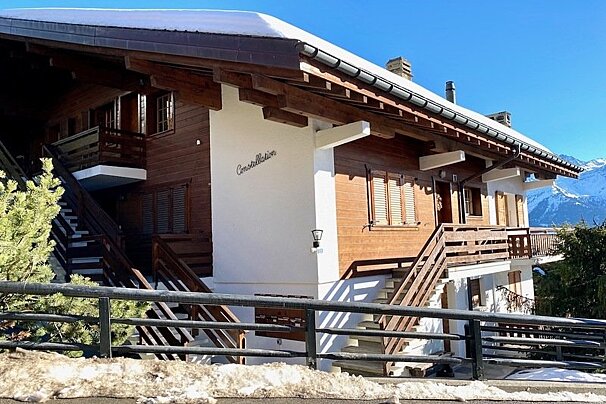 A wooden alpine chalet named "Constellation" with a snow-covered roof, set against a clear blue sky and distant snowy mountains.