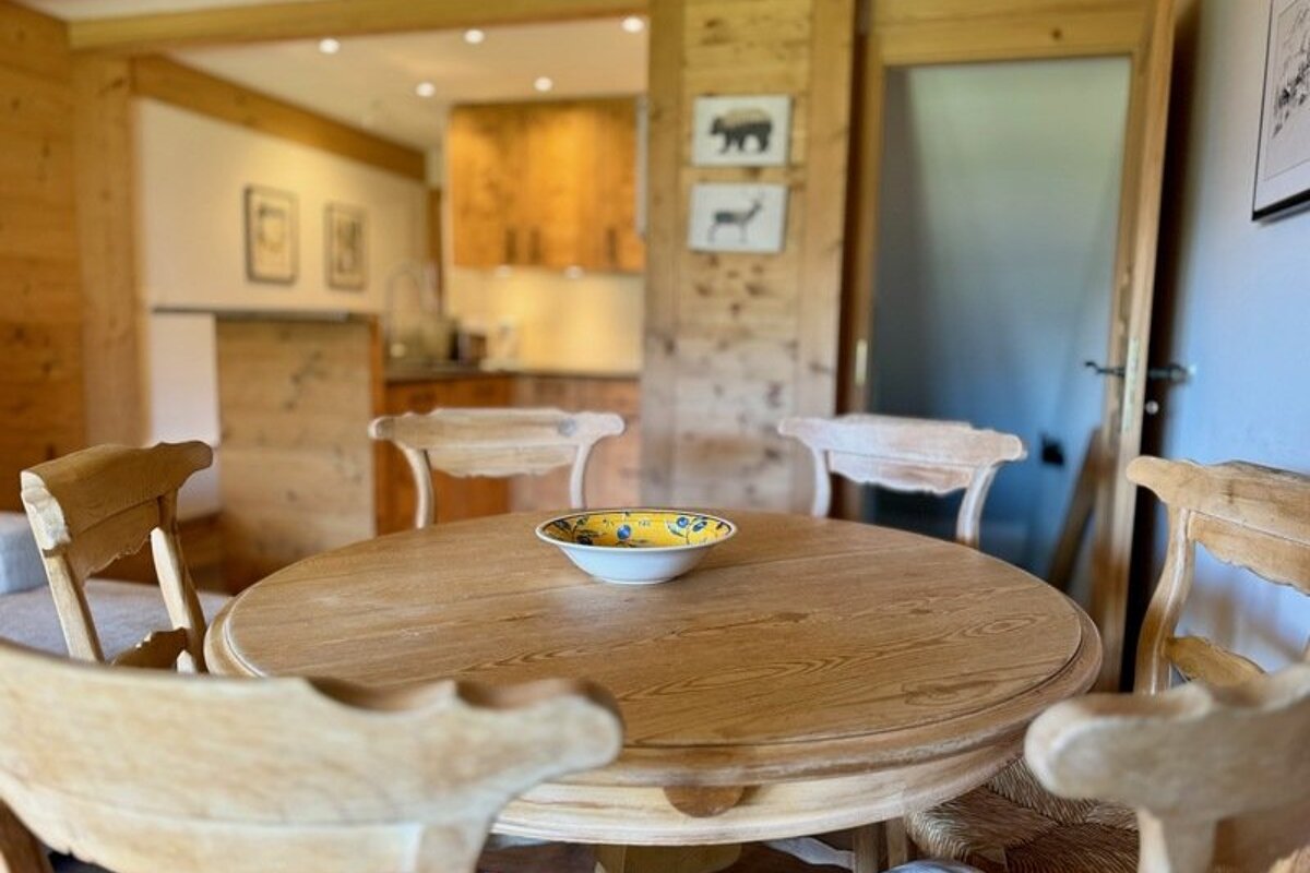 A round wooden dining table with a yellow bowl in the center, surrounded by wooden chairs in a cozy chalet-style home.