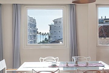 A bright dining room with a glass table and a window looking out at coastal buildings, palm trees, and the sea.