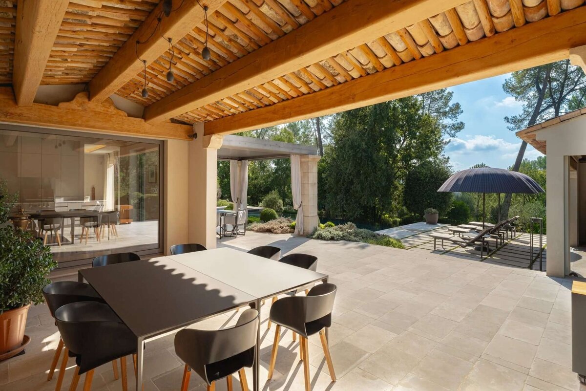 A spacious, sunlit outdoor patio with a dining area under a wooden beamed ceiling, overlooking a lush garden with lounge chairs and an umbrella.