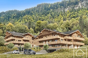 Two modern wooden alpine chalets nestled against a dense, green forested hillside with cars parked below.