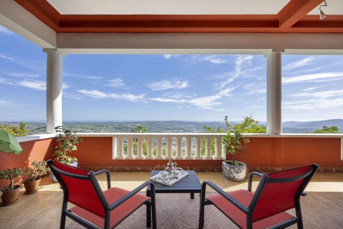 A scenic balcony features red chairs, a table with a tea set, and potted plants, overlooking a vast, green landscape under a bright blue, cloudy sky.