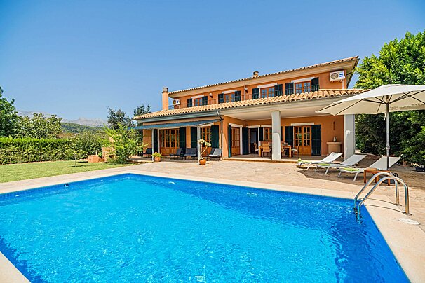 A vibrant orange villa with a sparkling blue pool, set against a backdrop of green trees and mountains under a clear sky.