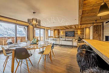 Spacious, luxurious chalet interior with warm wood accents, featuring a dining area, lounge, and large windows with stunning snowy mountain views.