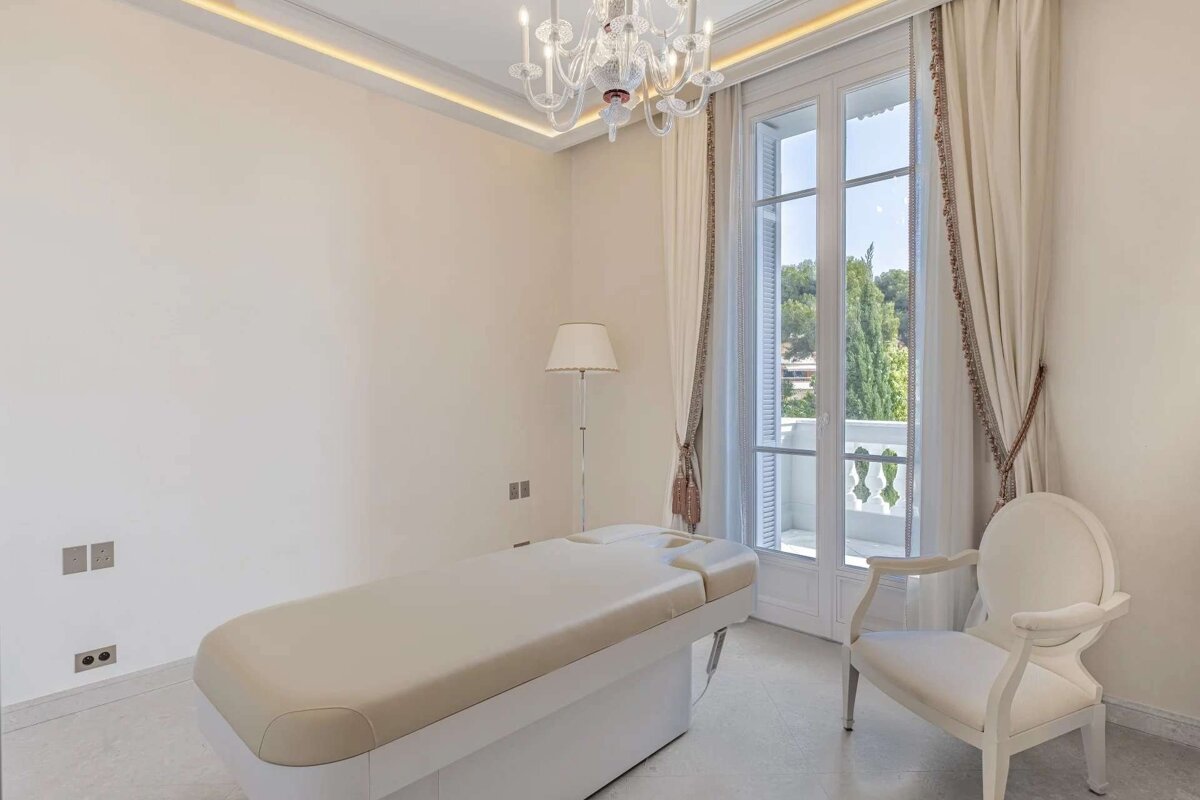 A bright, clean, spa-like room features a treatment table, elegant white armchair, floor lamp, and a large balcony door. A chandelier adds a luxurious touch.