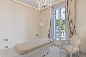 A bright, clean, spa-like room features a treatment table, elegant white armchair, floor lamp, and a large balcony door. A chandelier adds a luxurious touch.