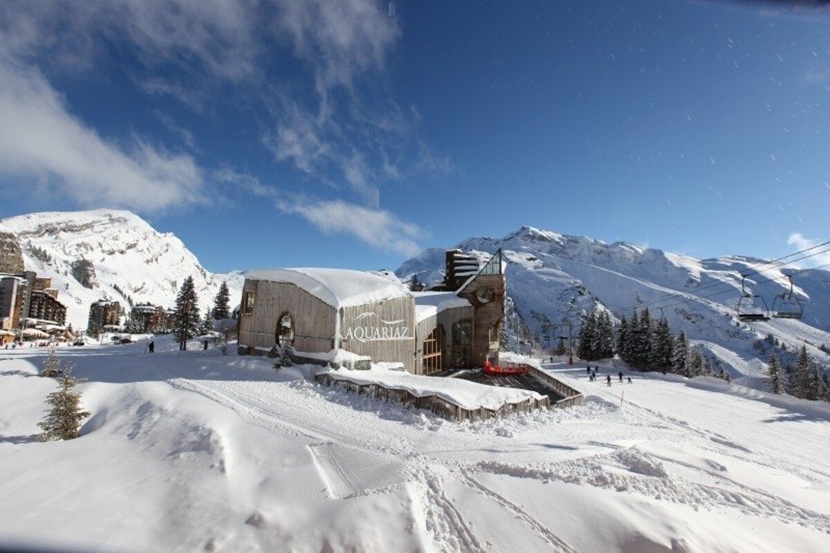 A snow-covered Aquariaz building at a sunny ski resort, featuring majestic mountains, ski lifts, and skiers under a clear blue sky.