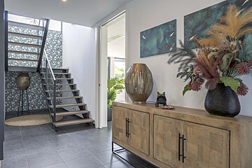 A modern entryway with a sleek wooden staircase and floral wallpaper. A wooden sideboard holds a striking lamp, a large dried flower arrangement, and abstract art.