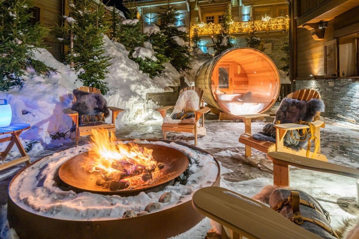 A cozy, snowy winter patio at night featuring a blazing fire pit, Adirondack chairs with fur throws, and a glowing barrel sauna/hot tub.