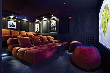 A luxurious home cinema with tiered plush red seating, a large screen displaying the Warner Bros. logo, and dark, paneled walls adorned with framed art.