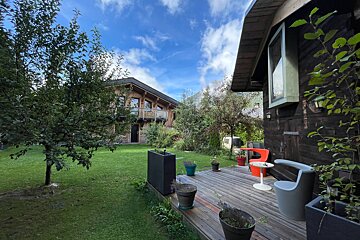 Two wooden chalets are surrounded by a lush green lawn and trees, with a deck and outdoor furniture in the foreground, under a blue, cloudy sky.