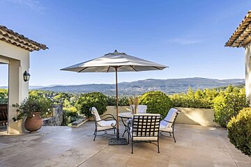 A sunny outdoor terrace with dining furniture and an umbrella, boasting panoramic views of a lush valley and distant mountains.