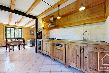 A rustic kitchen and dining area featuring natural wood cabinetry, exposed wooden beams, and tiled floors. A large window overlooks greenery.