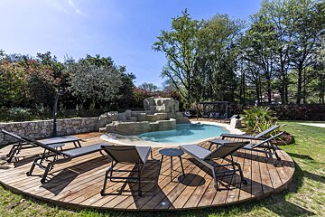 A sunny outdoor pool with a rock waterfall, surrounded by a round wooden deck with sun loungers and lush greenery.