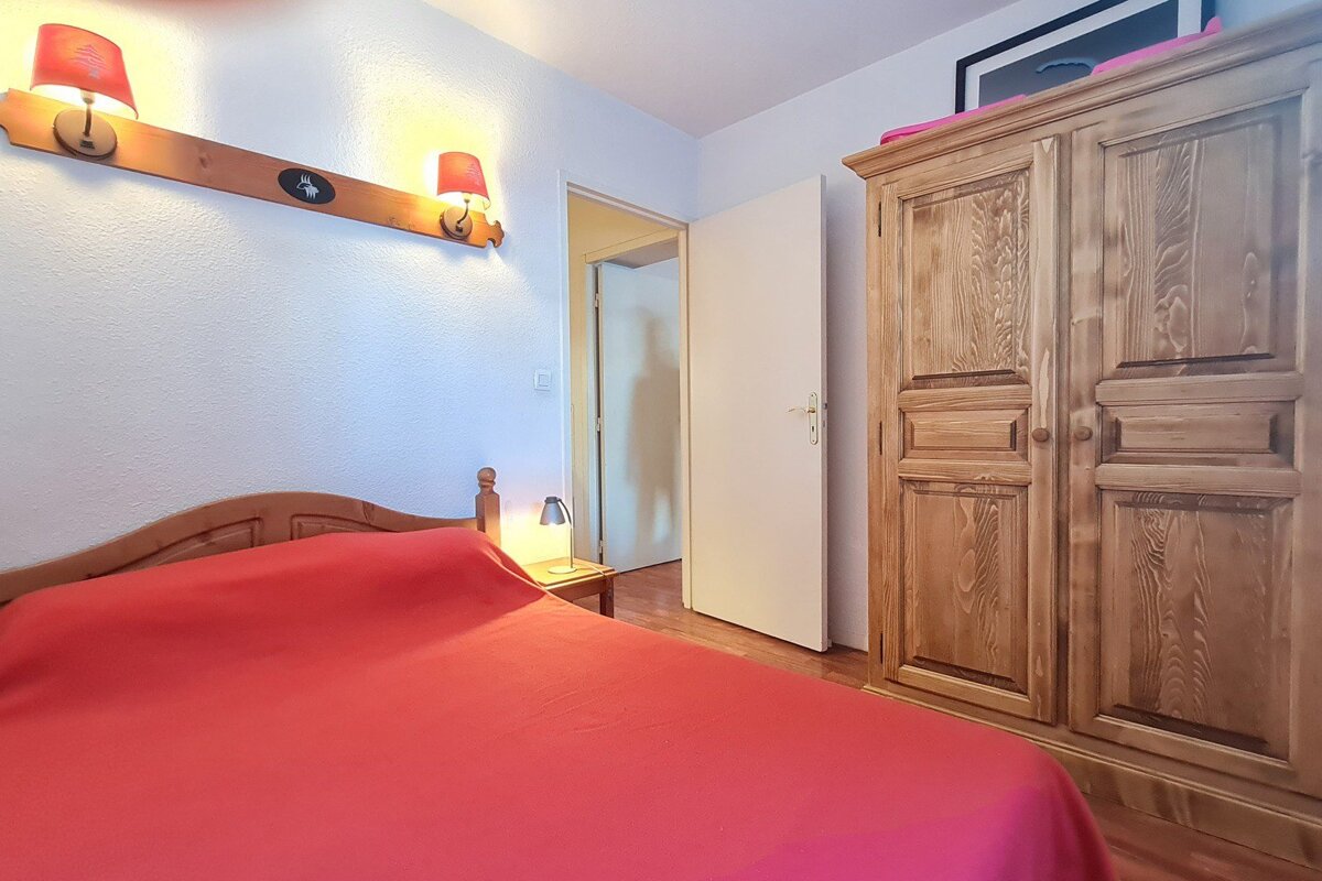 A cozy bedroom featuring a vibrant red bed, wooden headboard, rustic wooden wardrobe, and wall-mounted red-shaded lamps, with an open door visible.