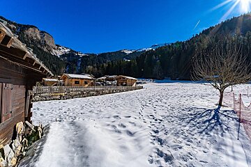 A snowy field with footprints in front of wooden chalets and forested mountains under a bright blue winter sky.