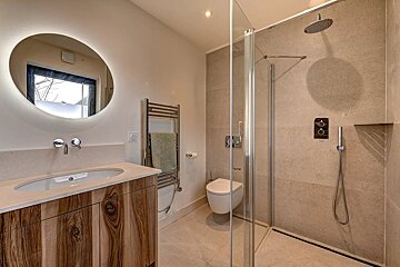 A stylish, contemporary bathroom featuring a walk-in shower, round backlit mirror, wood-grain vanity, and light tiled walls.