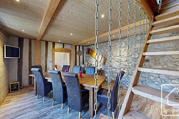 Rustic dining room with a long wooden table, plush chairs, exposed wood beams, a stone wall, and a wooden staircase with decorative chains.