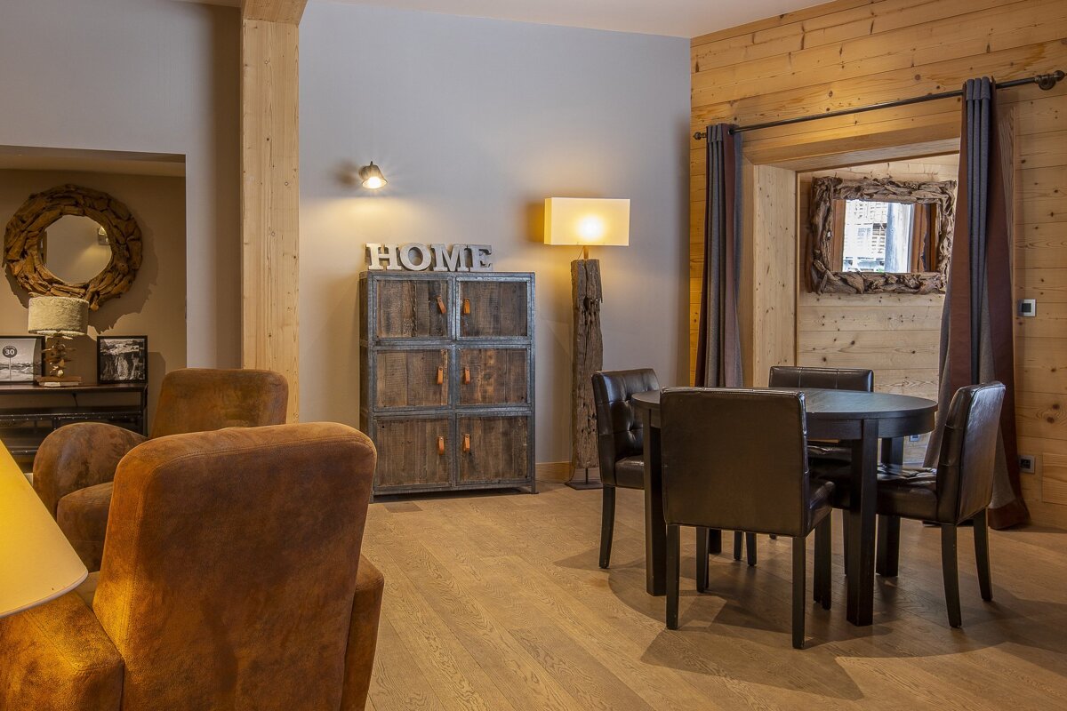 A warm, rustic living space featuring a round dining table, brown armchairs, wooden walls, and a 