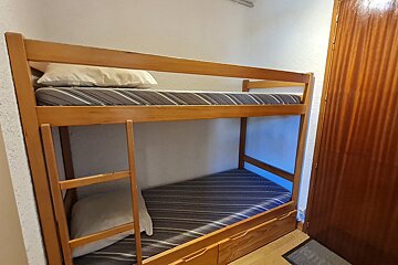 A light wooden bunk bed with two striped mattresses and pillows stands against a white wall. A dark wooden door is to the right.