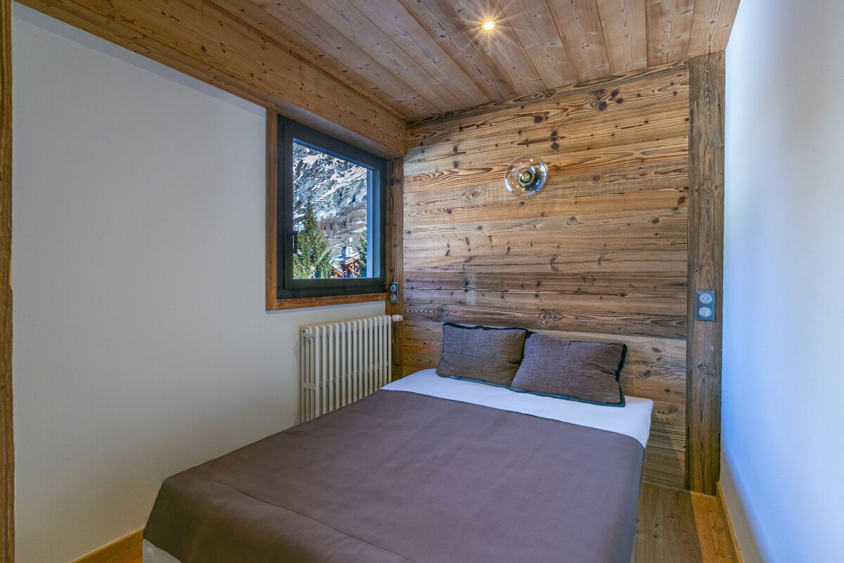 Cozy bedroom featuring wood-paneled walls and ceiling, a bed with a brown blanket, and a window with a view of snowy mountains.