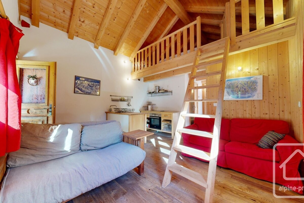 A cozy, wood-paneled chalet room with a sleeping loft accessed by a ladder, featuring a gray futon, red sofa, and compact kitchen area.