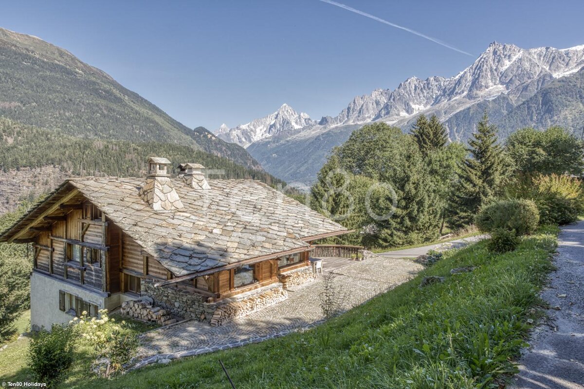 A house with a stone roof is surrounded by mountains and trees
