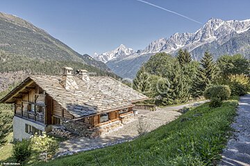 A house with a stone roof is surrounded by mountains and trees