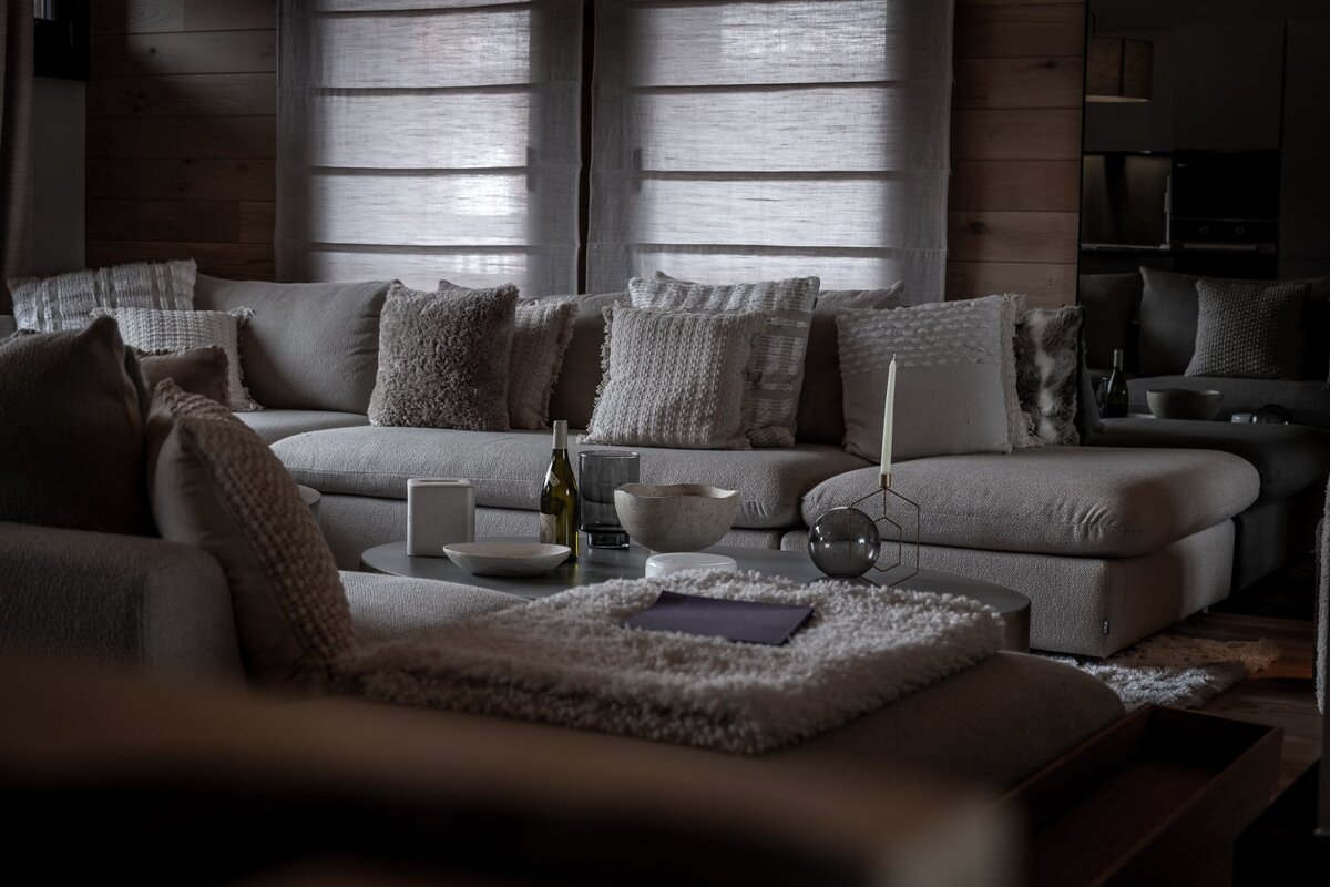 A cozy, dimly lit living room with a large grey sectional sofa, many textured pillows, and a coffee table with a wine bottle and candle.