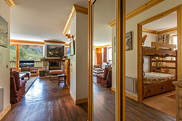 Cozy chalet living room with mountain views, a fireplace, and a mirrored hallway leading to a bedroom with rustic wooden bunk beds.