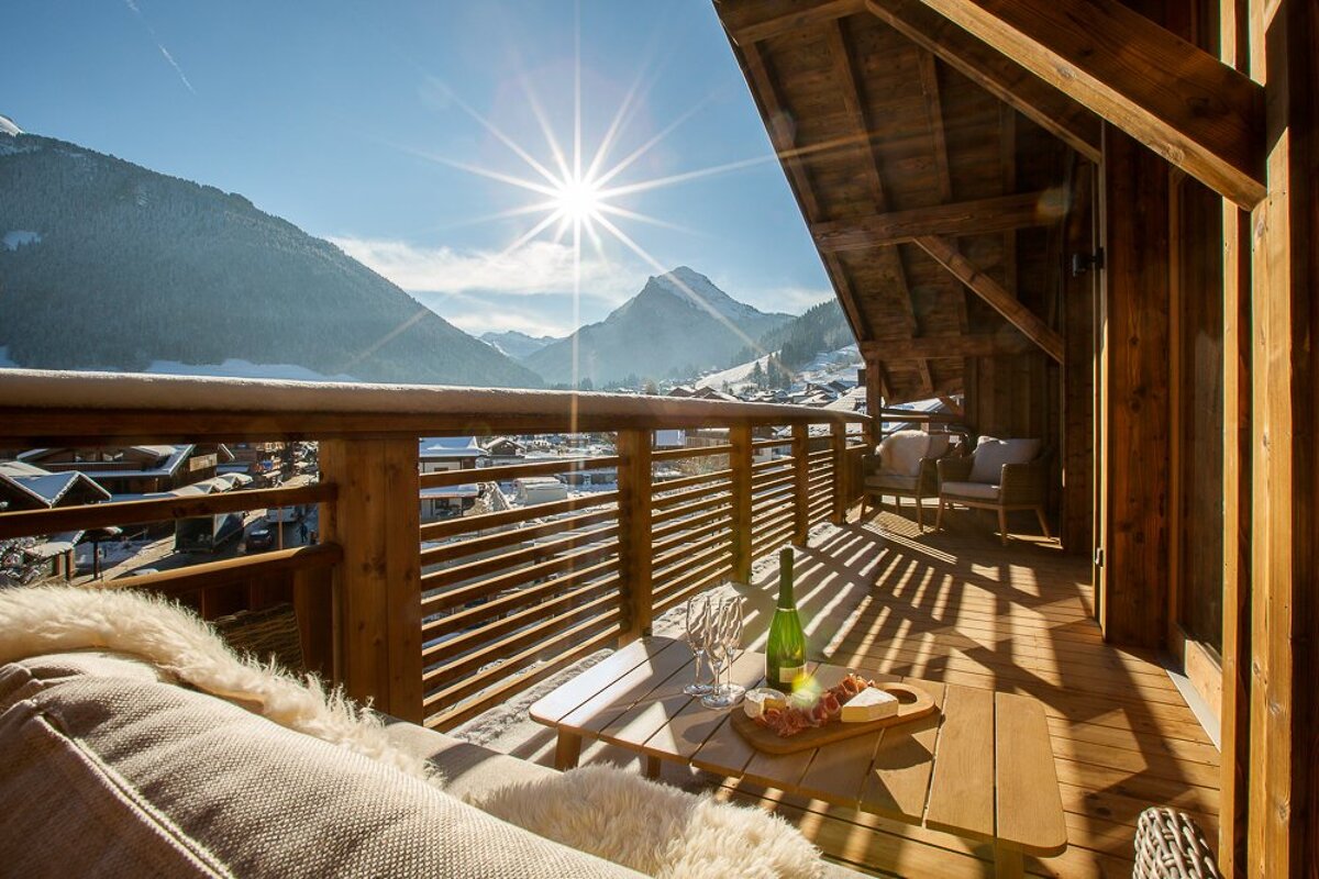 A sunny wooden balcony with champagne, cheese, and cozy seating offers a stunning view of a snowy mountain village and bright sky.