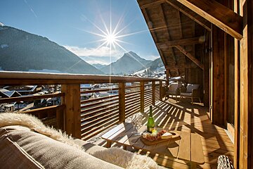 A sunny wooden balcony with champagne, cheese, and cozy seating offers a stunning view of a snowy mountain village and bright sky.