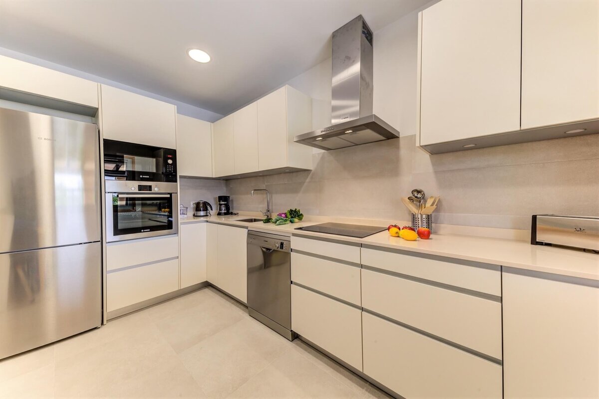 A bright, modern L-shaped kitchen with cream cabinets, stainless steel appliances (fridge, oven, microwave, dishwasher, hood), and light countertops.