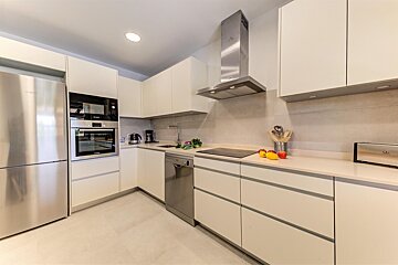 A bright, modern L-shaped kitchen with cream cabinets, stainless steel appliances (fridge, oven, microwave, dishwasher, hood), and light countertops.