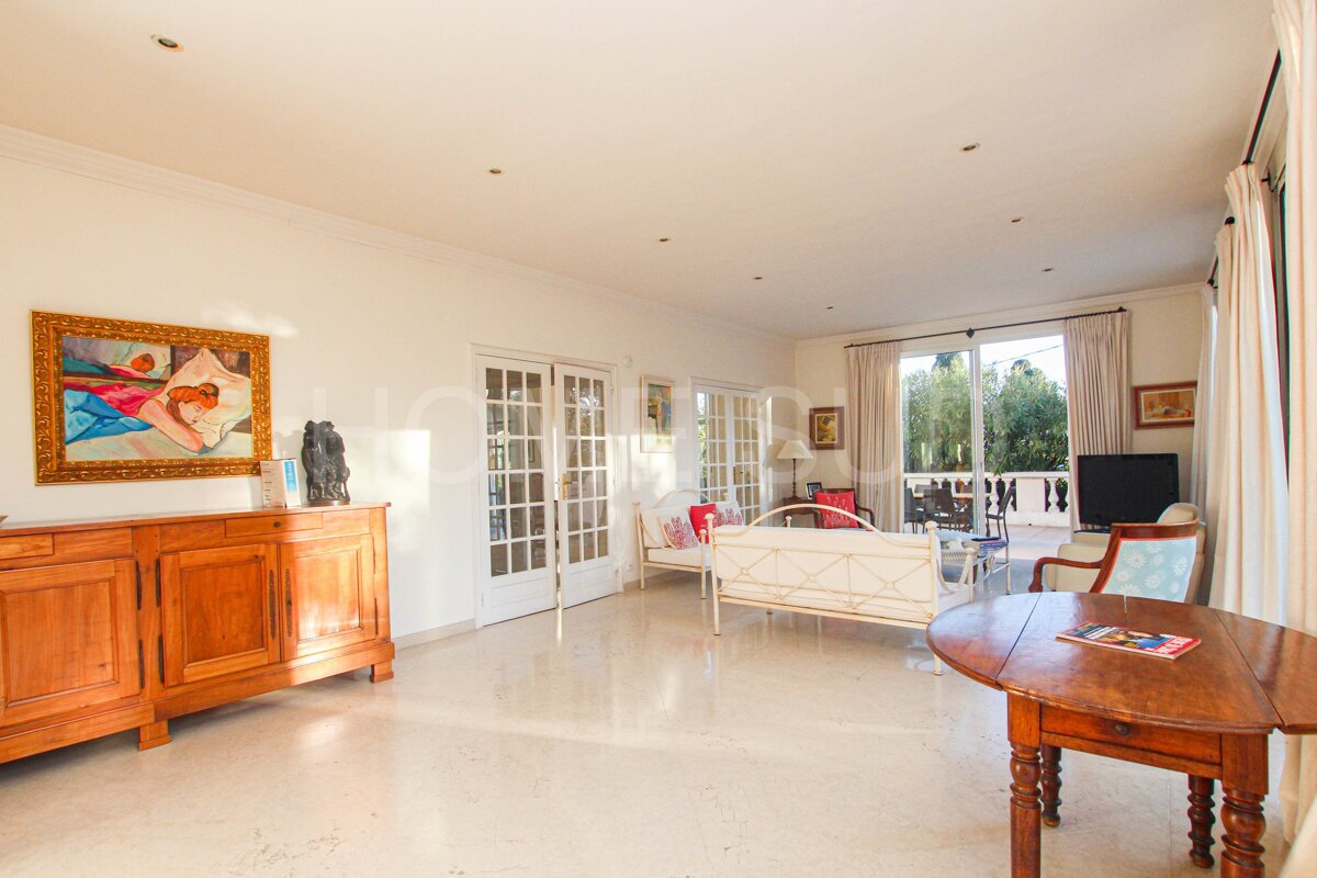 A sunlit, spacious living room with white walls, tiled floor, a wooden sideboard, and glass doors opening to an outdoor area.