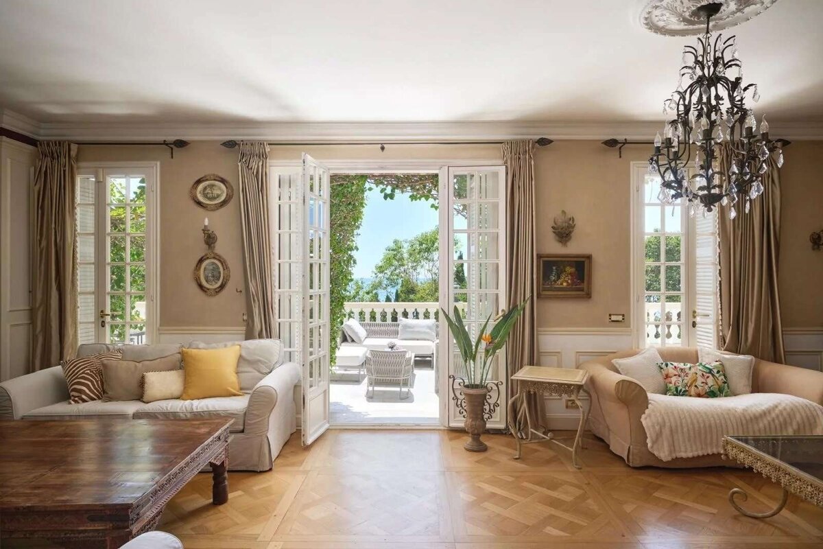 Elegant living room with French doors opening to a sunny terrace overlooking the sea, featuring luxurious sofas, a chandelier, and parquet flooring.