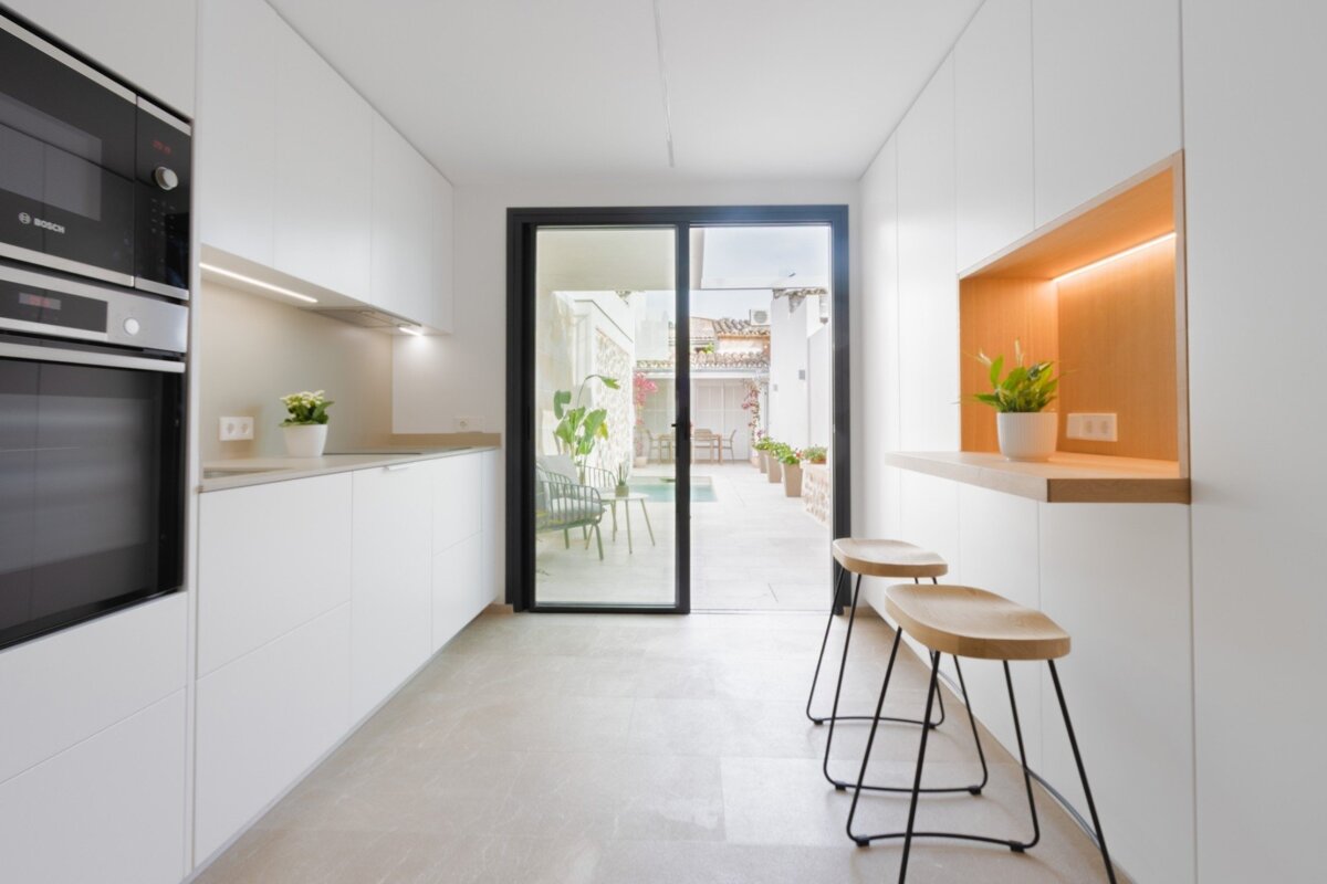 A modern, minimalist white kitchen features built-in appliances, a wood-accented bar with stools, and sliding glass doors leading to an outdoor patio.