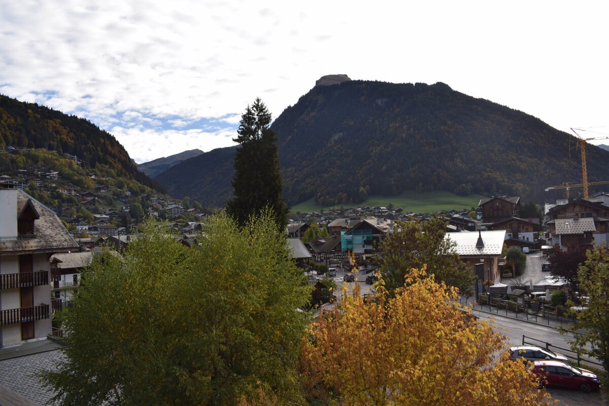 A picturesque mountain town nestled in a valley, with colorful autumn trees in the foreground and towering forested peaks behind.