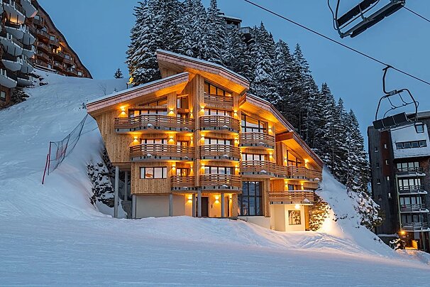 A brightly lit modern wooden chalet is nestled in a snowy mountain landscape with pine trees and ski lifts under a dark sky.
