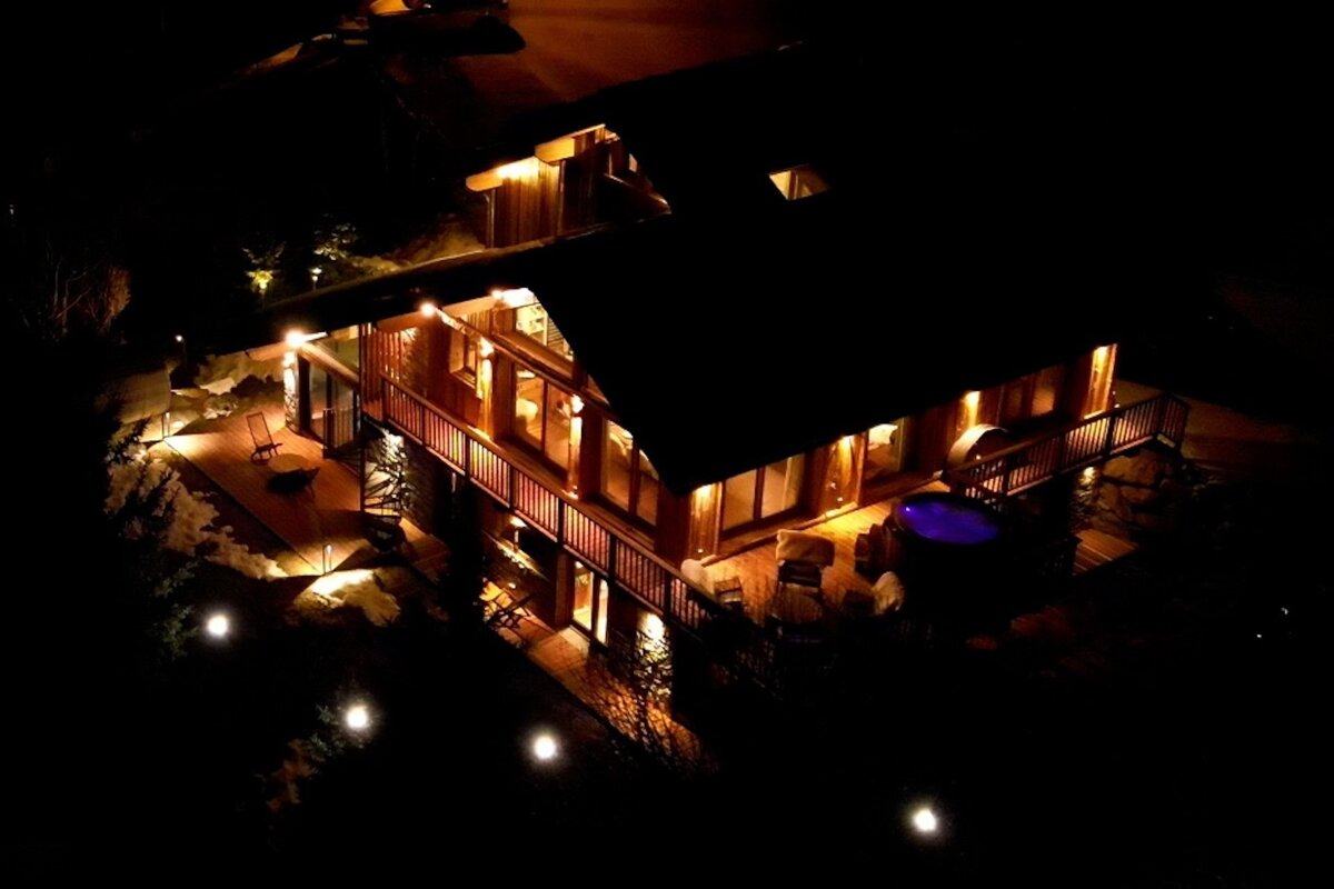 A cozy, warmly lit wooden lodge at night, featuring spacious decks, glowing interiors, and a vibrant blue hot tub under the dark sky.