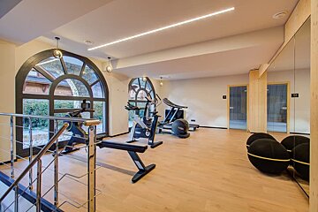 A modern gym room with wooden flooring, various fitness equipment, large arched windows, and a mirrored wall.